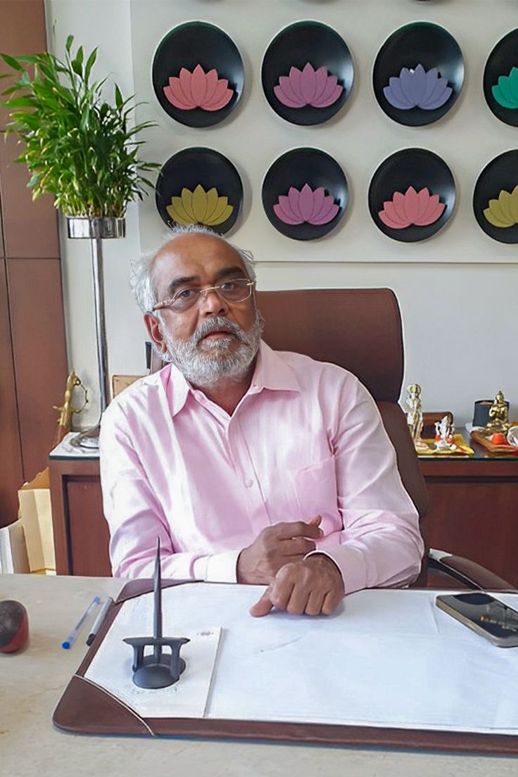 Savji Dholakia seated at his office desk in a pink shirt, with decorative lotus plates on the wall behind him.