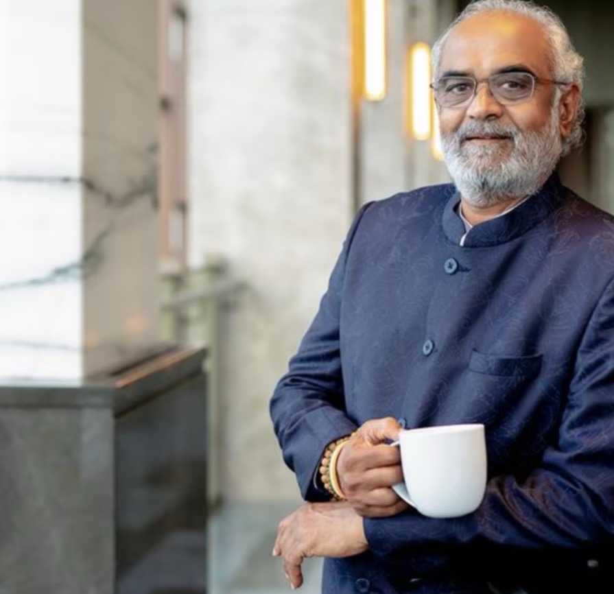 Savji Dholakia standing by a window holding a white cup, wearing a dark indigo bandhgala jacket.