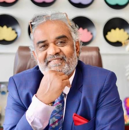 Savji Dholakia resting his chin on his hand in his office, wearing a blue checked suit.