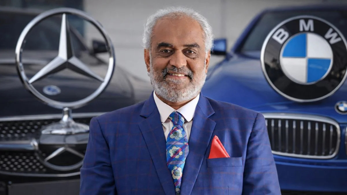 Savji Dholakia photographed in a blue suit standing in front of two cars.