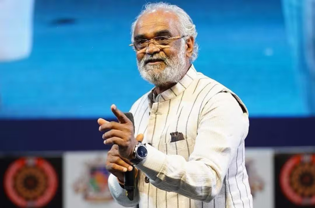 Savji Dholakia speaking on stage with a microphone in his hand.