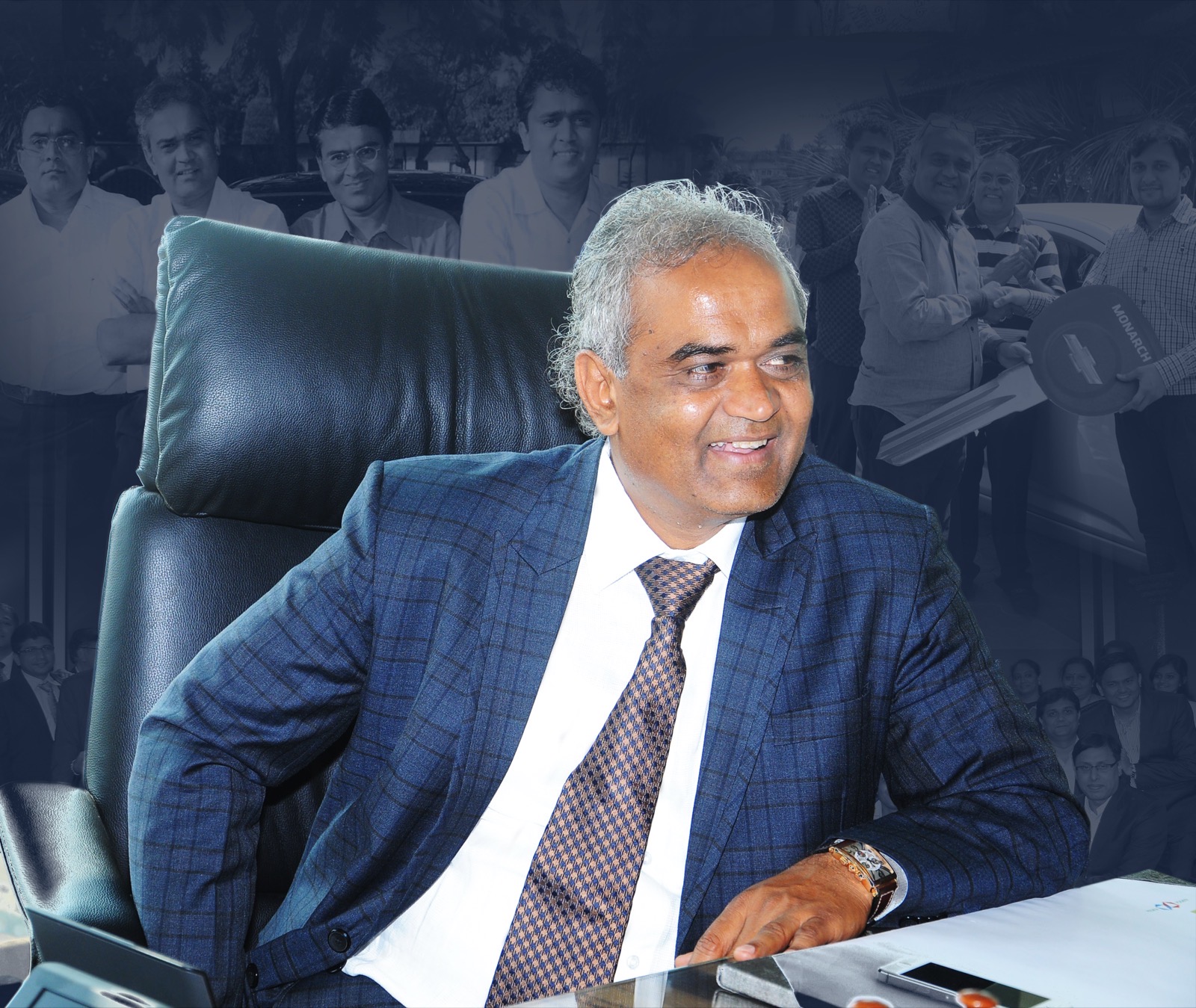 Savji Dholakia seated at his desk in a blue suit, with archive photographs of company events behind him.