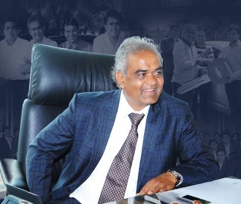 Savji Dholakia seated at his desk in a blue suit, smiling and looking to one side.
