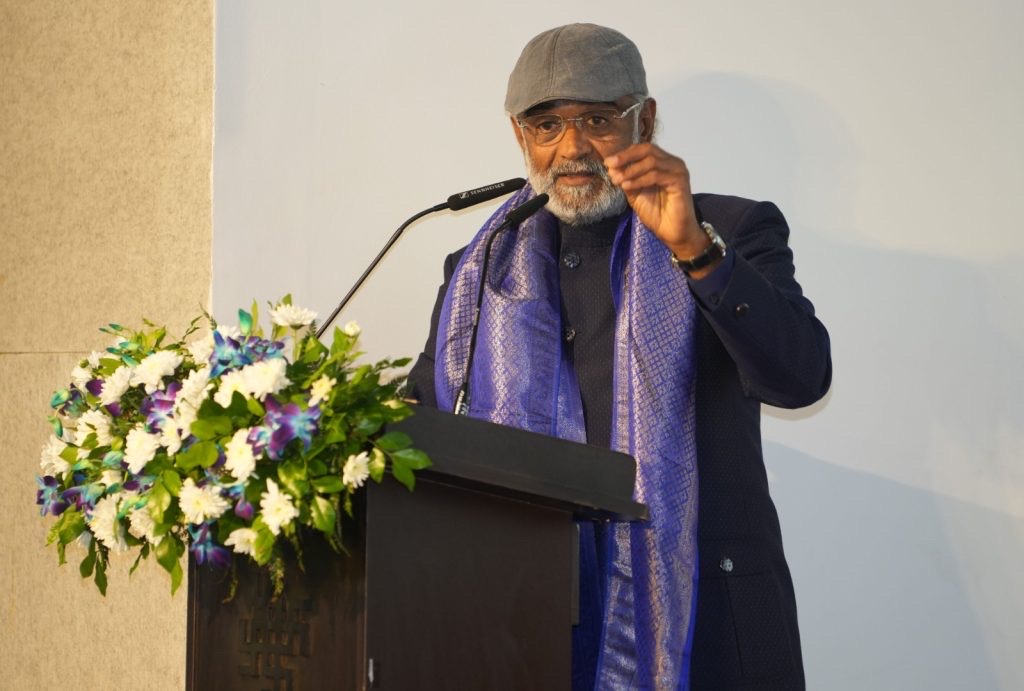 Savji Dholakia speaking from a lectern, wearing a cap and a purple shawl.
