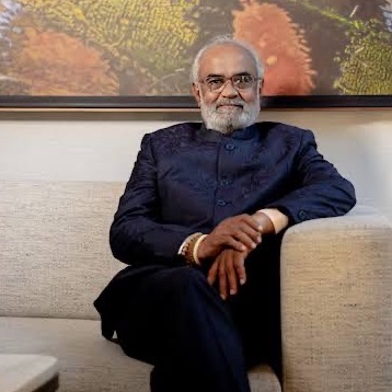 Savji Dholakia seated on a sofa in a dark indigo bandhgala, hands resting together.